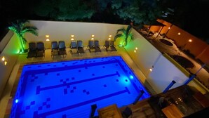Una piscina con un deck iluminado en azul, rodeada de sillas de descanso y palmeras.