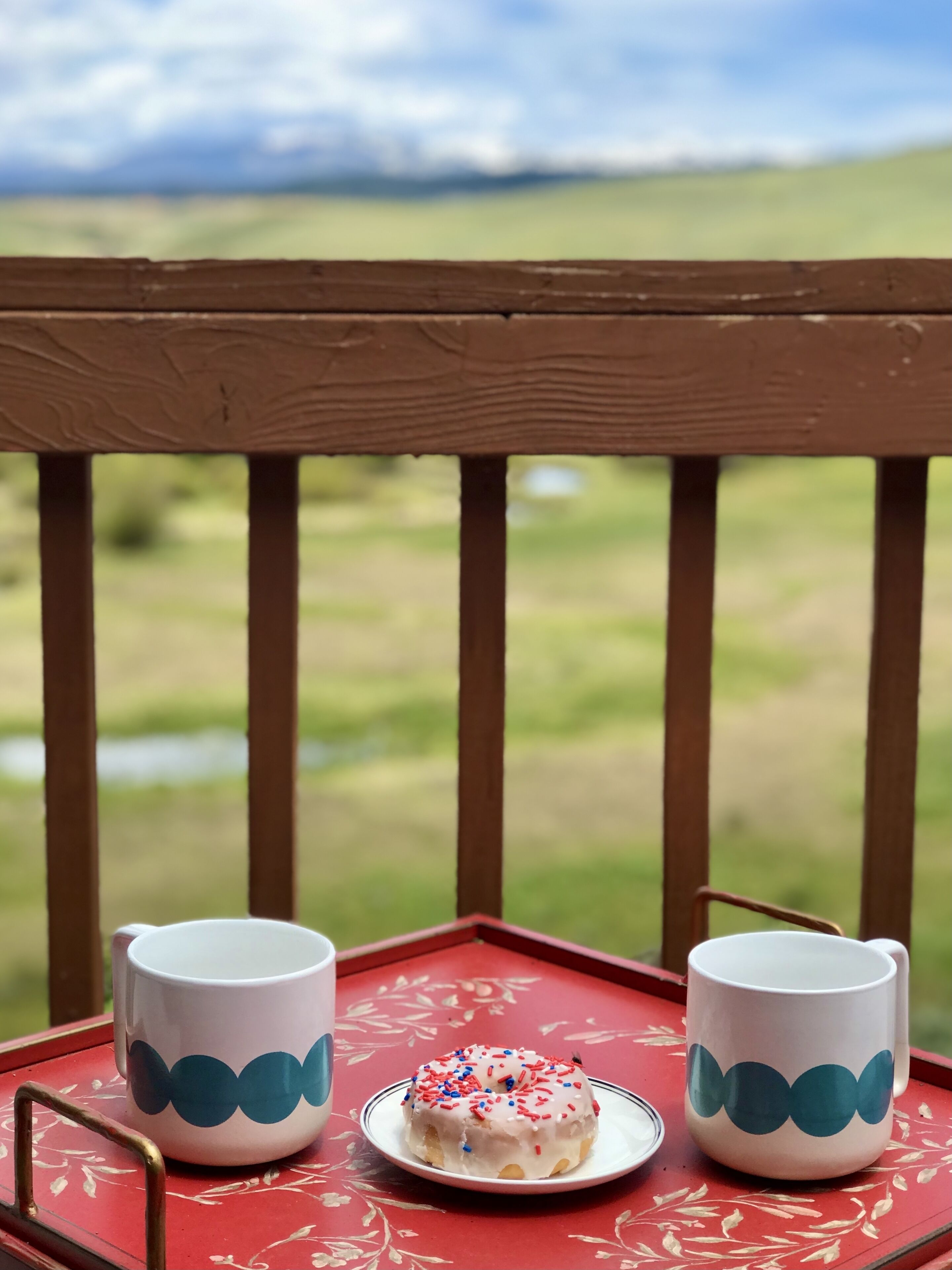 Une table rouge avec deux tasses blanches et une assiette sur laquelle se trouve un beignet, le tout sur un balcon donnant sur un paysage verdoyant.