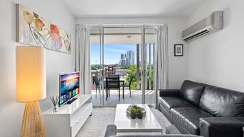 A modern living room with a flat-screen TV, a black leather sofa, a coffee table, and a dining area with a table and chairs.