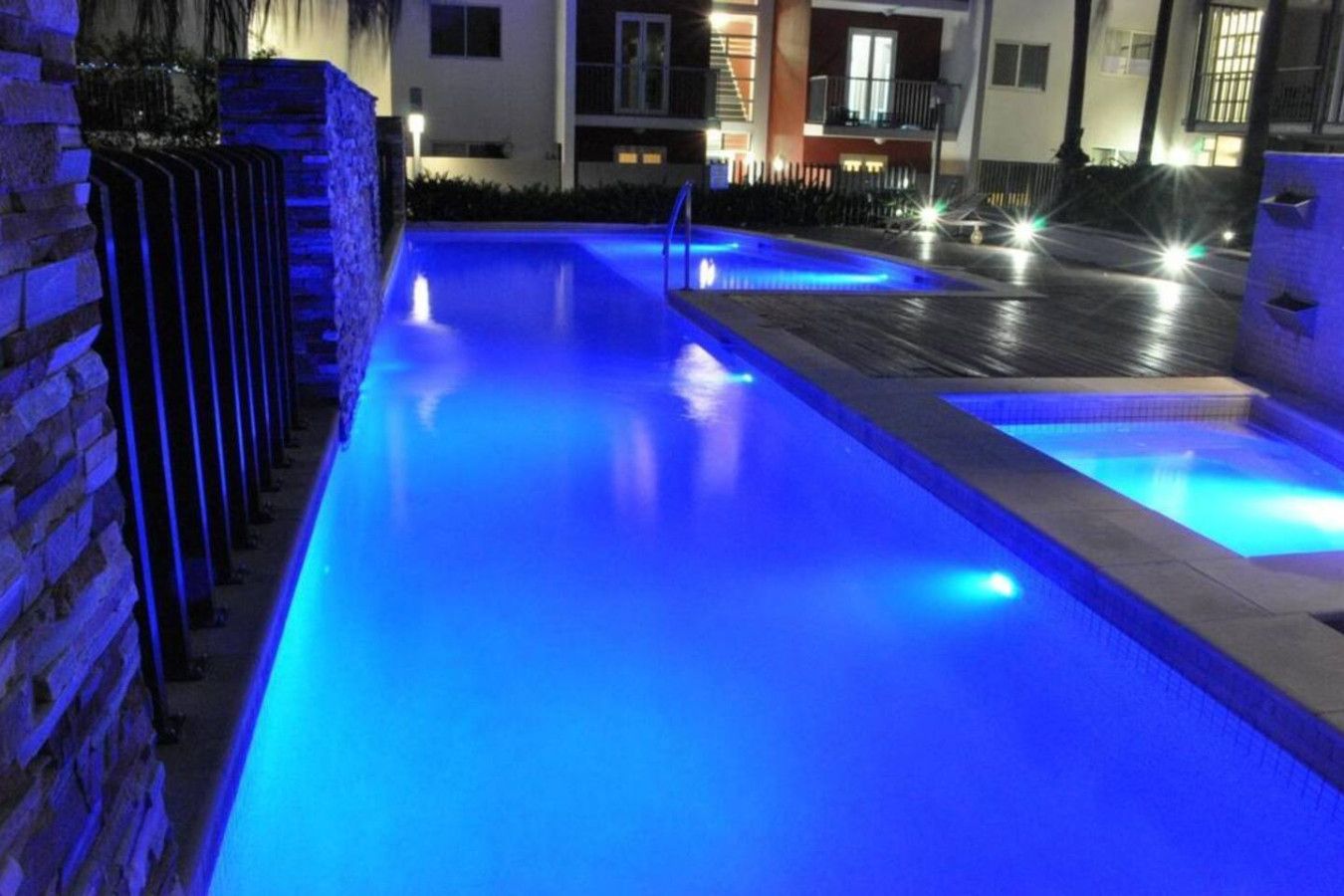 A swimming pool with blue lighting, surrounded by a brick wall and a building with illuminated windows.