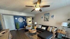 A modern living room with a black sofa, a wooden coffee table, and a ceiling fan. There is a door leading to a kitchen and a dining area.