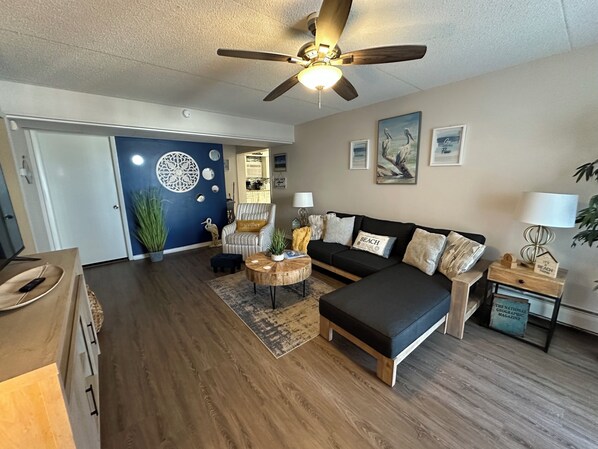 A modern living room with a black sofa, a wooden coffee table, and a ceiling fan. There is a door leading to a kitchen and a dining area.