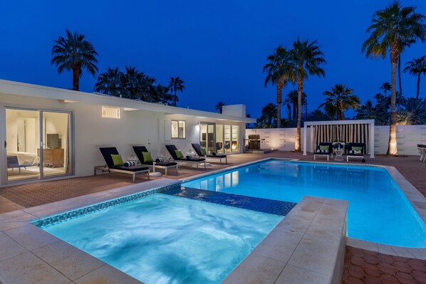 Un área de piscina moderna con sillones de descanso, una alberca y palmeras.
