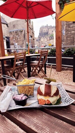 Un repas composé d’un sandwich, de frites et d’une boisson, servi sur une table en bois sous un parasol rouge, avec une vue sur la ville en arrière-plan.
