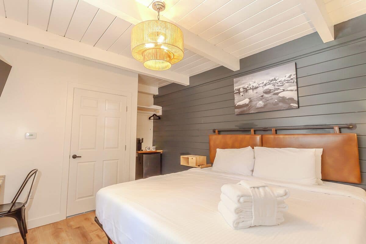 A neatly arranged bedroom with a bed, a hanging light fixture, a painting on the wall, and a small wooden side table.