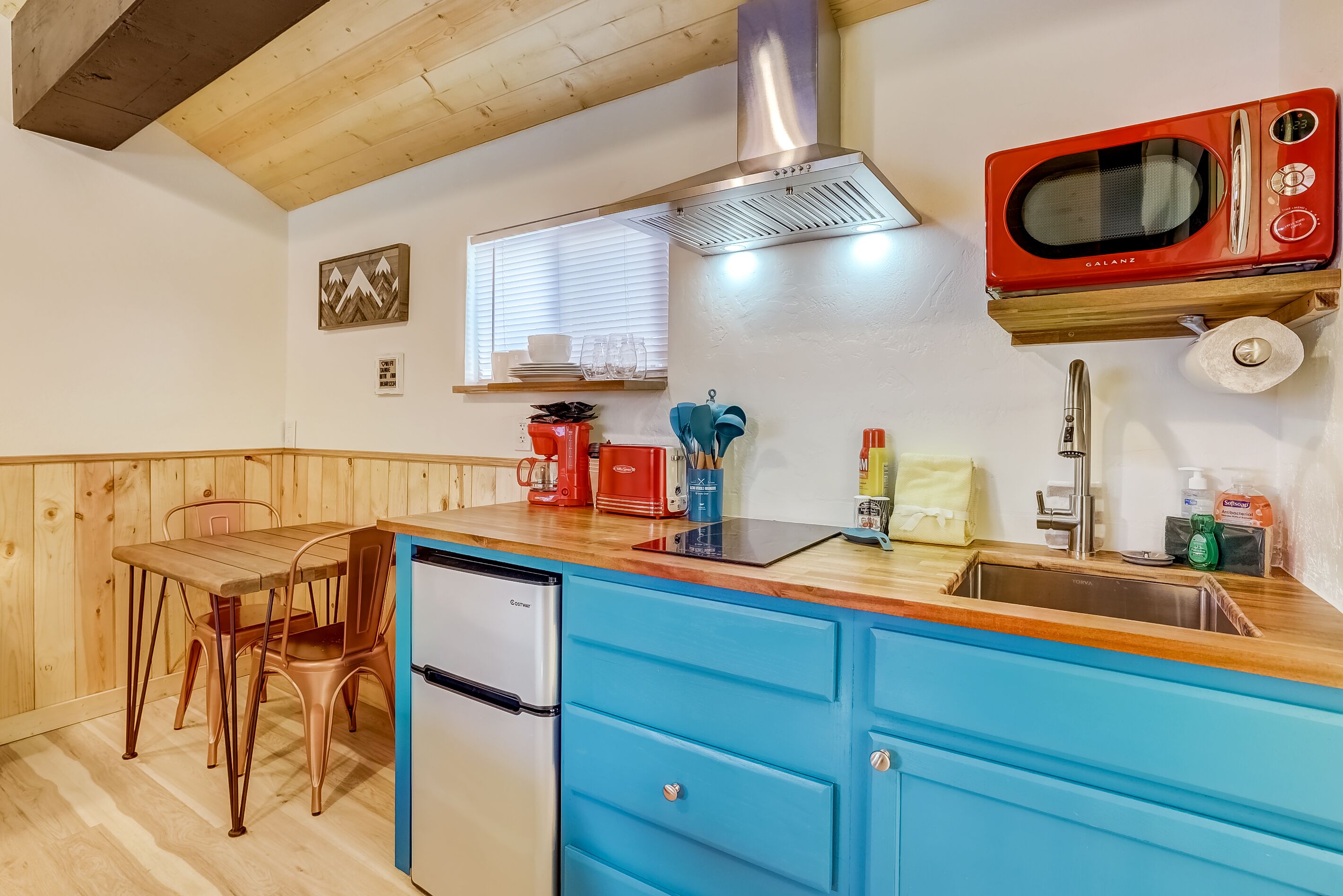Cocina compacta con armarios de madera, un refrigerador de acero inoxidable, un microondas rojo y una ventana con persianas.