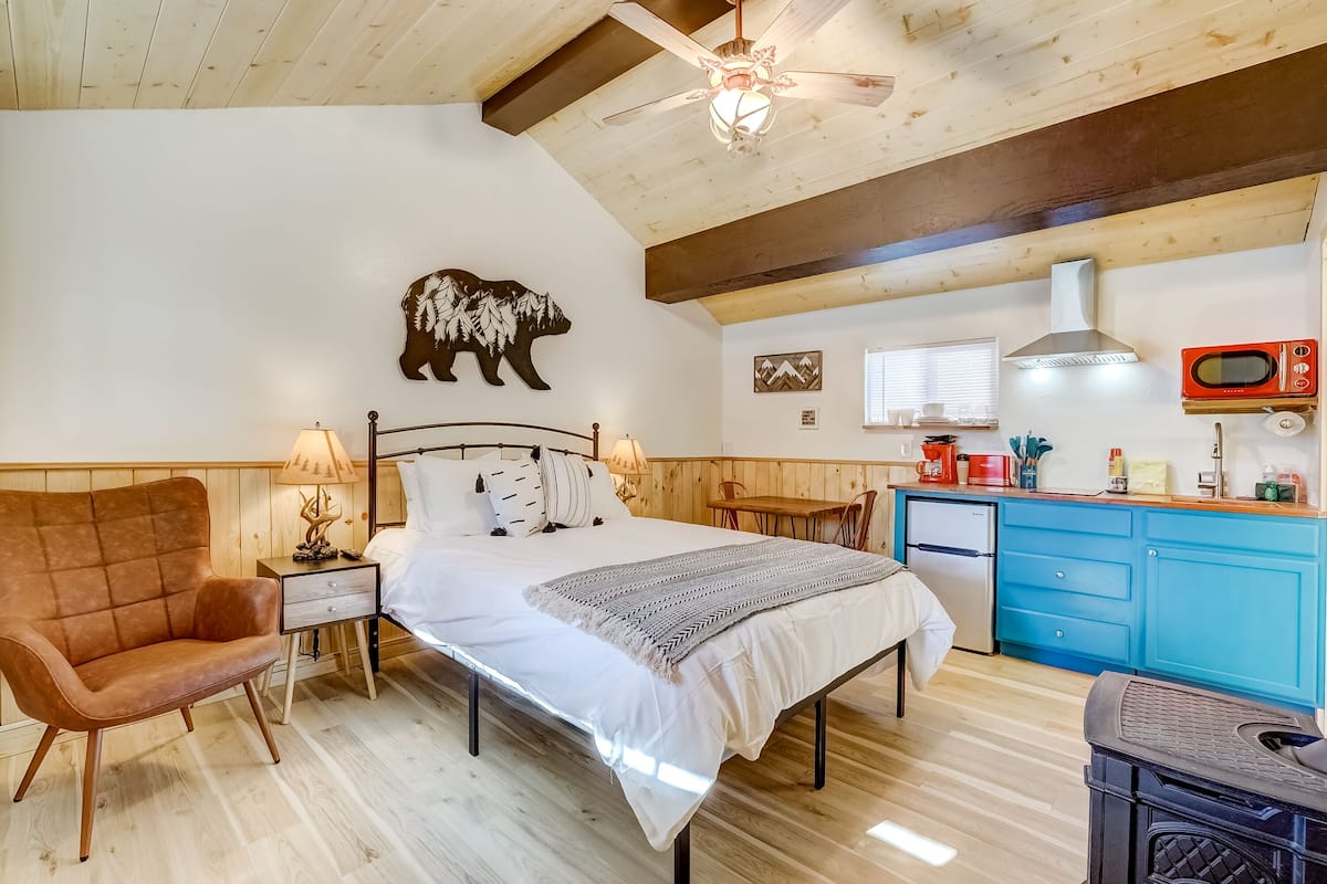 A bedroom with a bed, a nightstand, a chair, a wooden ceiling, a bear wall art, a ceiling fan, a kitchenette with a microwave, and a stove.