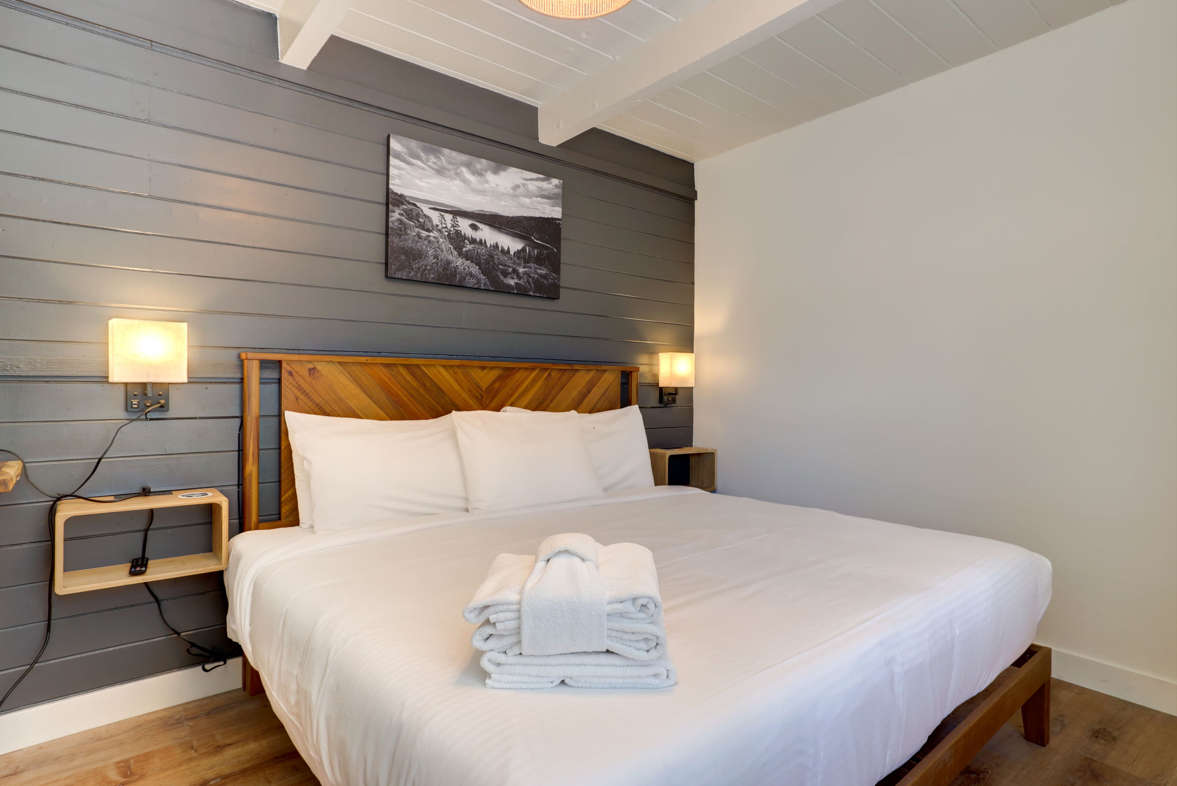 Un lit bien fait, avec du linge de lit blanc et une serviette pliée, une tête de lit en bois, des lampes de chevet et un tableau encadré au mur.