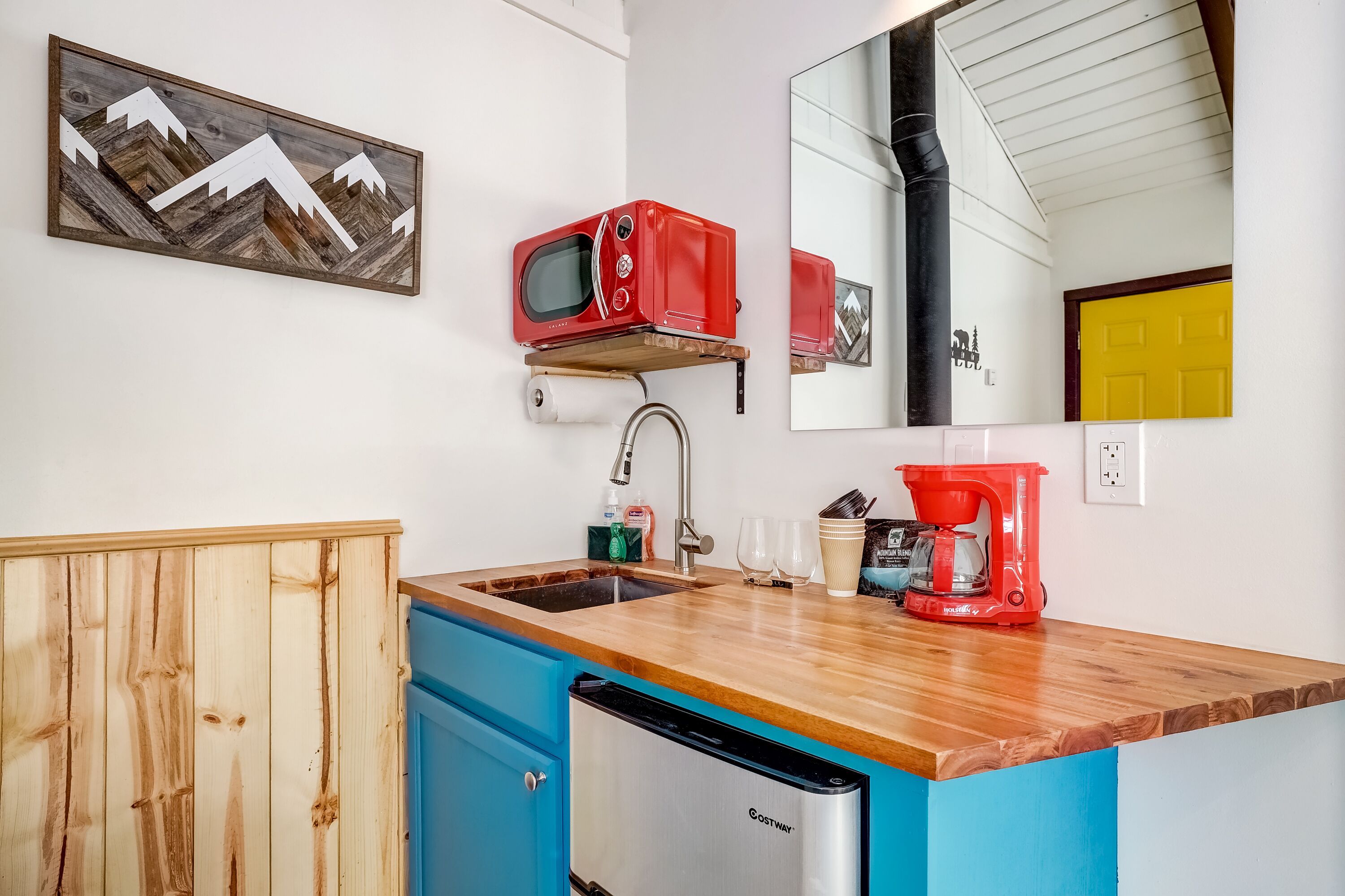 Cocina compacta con encimera de madera, un lavamanos, una cafetera roja y un microondas en un estante.