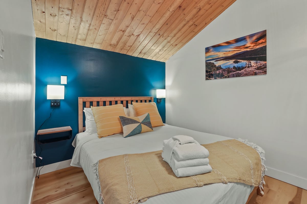 A bedroom with a wooden ceiling, a blue accent wall, a bed with white bedding, a yellow throw blanket, and a painting of a landscape.