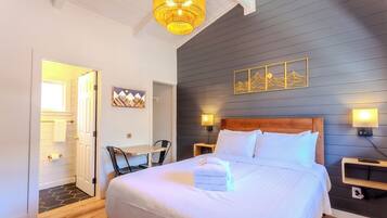 Un dormitorio con una cama grande, mesitas de noche, un escritorio y una silla. Hay una puerta que da a otra habitación.