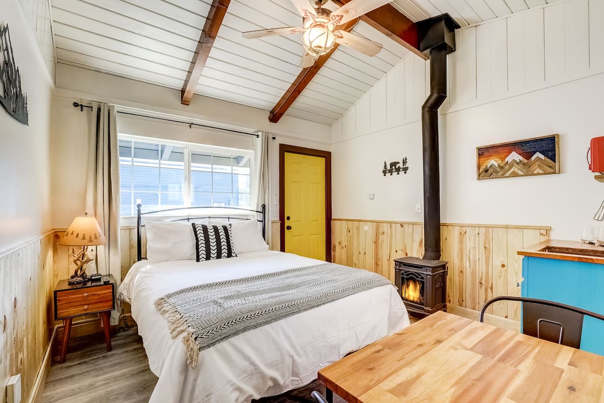 A cozy bedroom with a bed, bedside table, lamp, and a wood-burning stove.