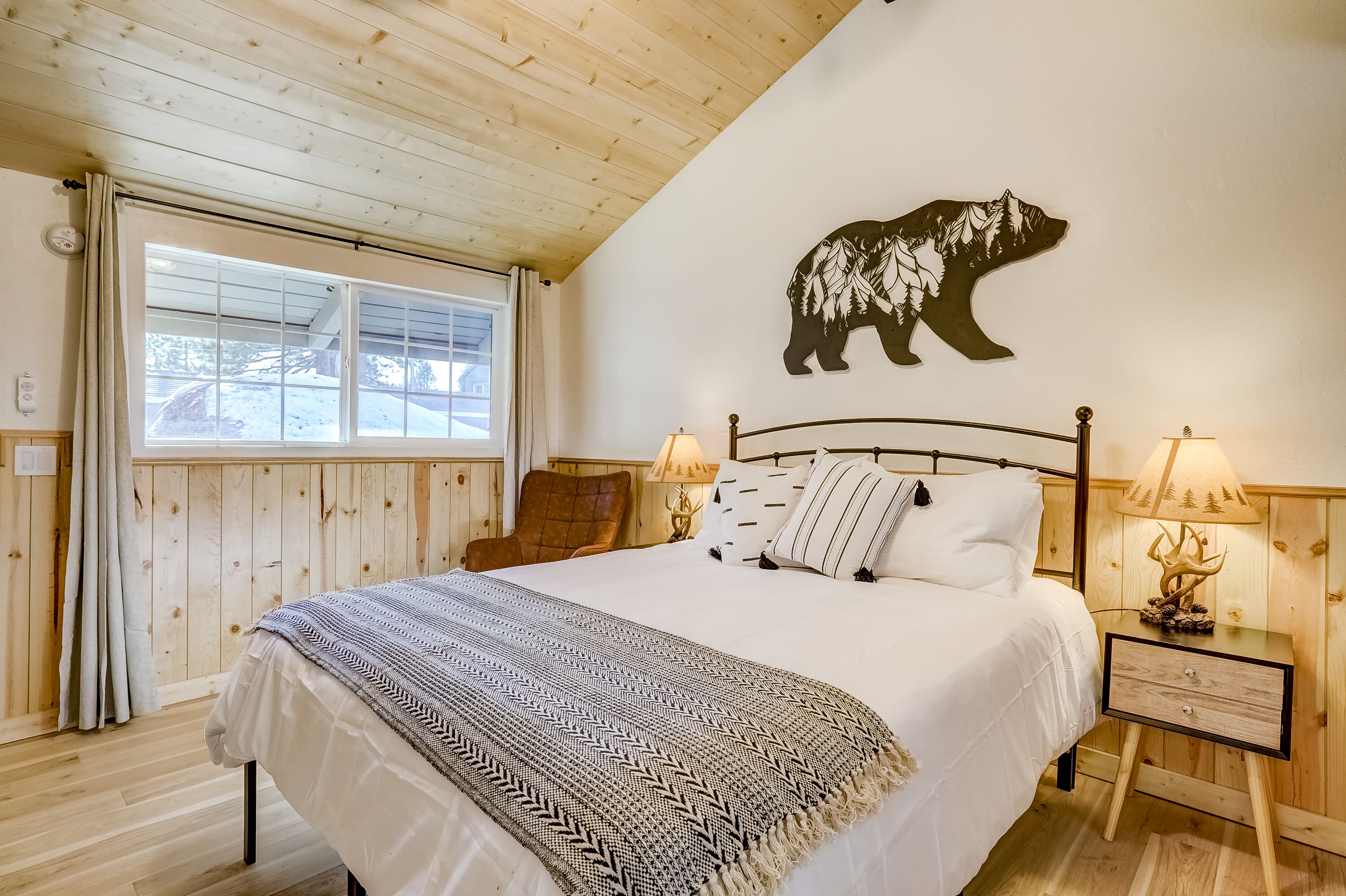 Un dormitorio acogedor con una cama grande, paredes de madera, un cuadro de un oso, dos lámparas de noche y una ventana con cortinas.