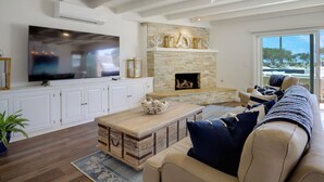 A living room with a stone fireplace, a large flat-screen TV, white cabinetry, and a beige sofa with blue cushions.
