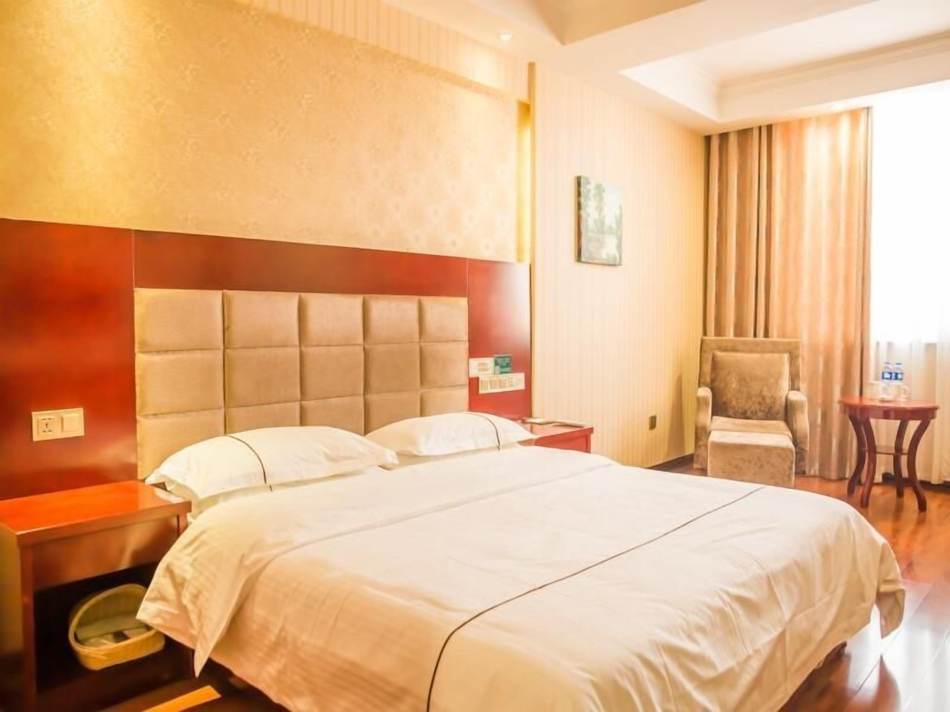 A hotel room with a large bed, a wooden nightstand, a chair, and a small table with a bottle.