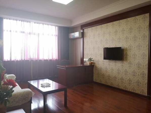 A living room with a wooden floor, a glass coffee table, a sofa, a TV mounted on the wall, and a window with curtains. - Greentree Inn Jiayuguan Xinhua Nan Road (Jiayuguan)