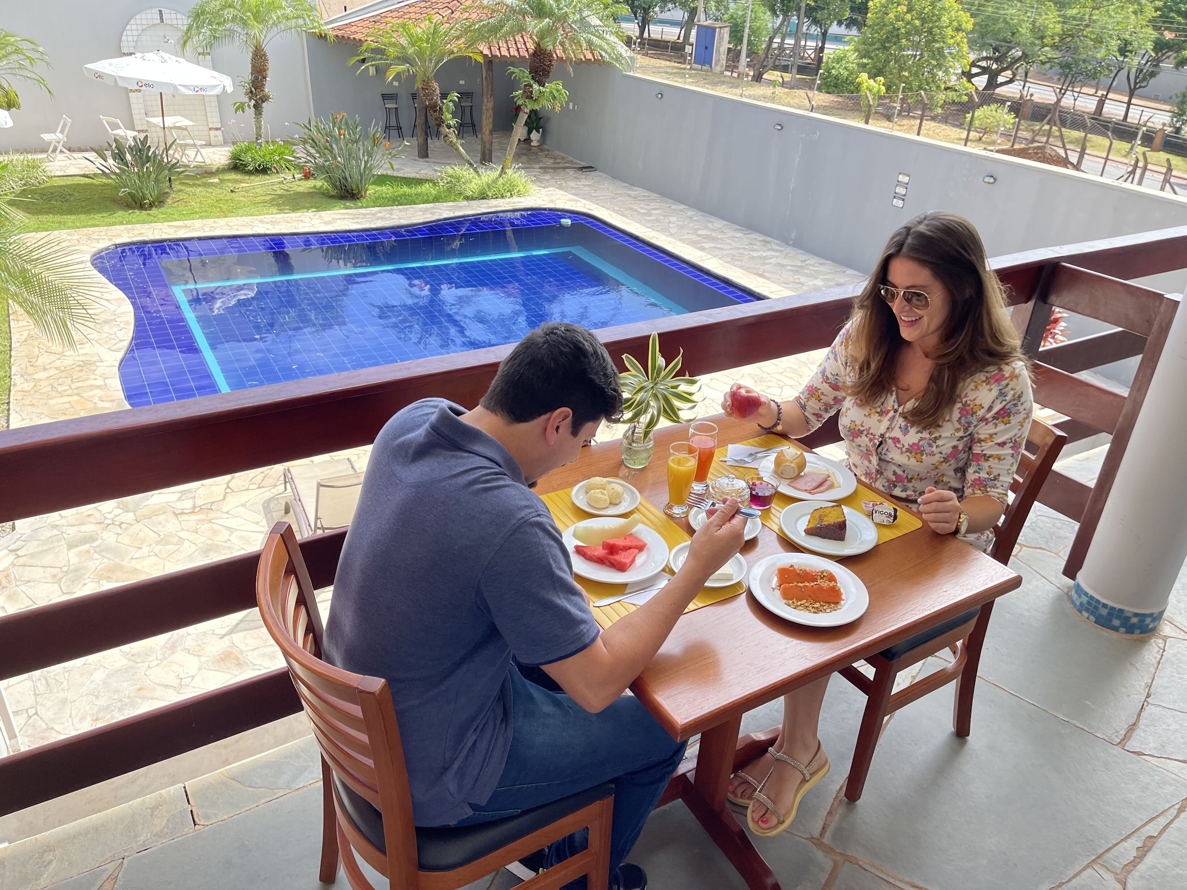 Un homme et une femme dînent à une table en bois, avec vue sur une piscine et un espace extérieur aménagé pour se détendre.