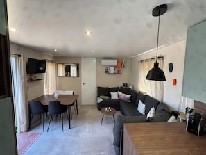 A modern living room with a sofa, a dining table with chairs, a TV mounted on the wall, and a ceiling light.