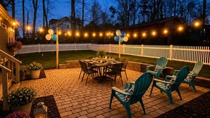 Un patio al aire libre con piso de ladrillo, una mesa de comedor, sillas, luces colgantes y una cerca de piquetes blancos.