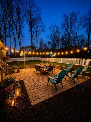 An outdoor patio with a brick floor, a dining table, chairs, string lights, and a white picket fence.