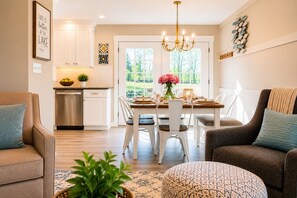 A modern kitchen with white cabinets, a dining area with a wooden table and chairs, and a view of greenery outside.
