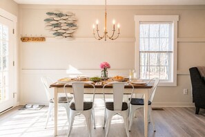 A dining area with a wooden table, white chairs, a chandelier, and a window with blinds.