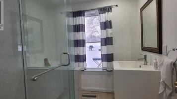 A bathroom with a glass shower door, a window with striped curtains, a sink, and a mirror.