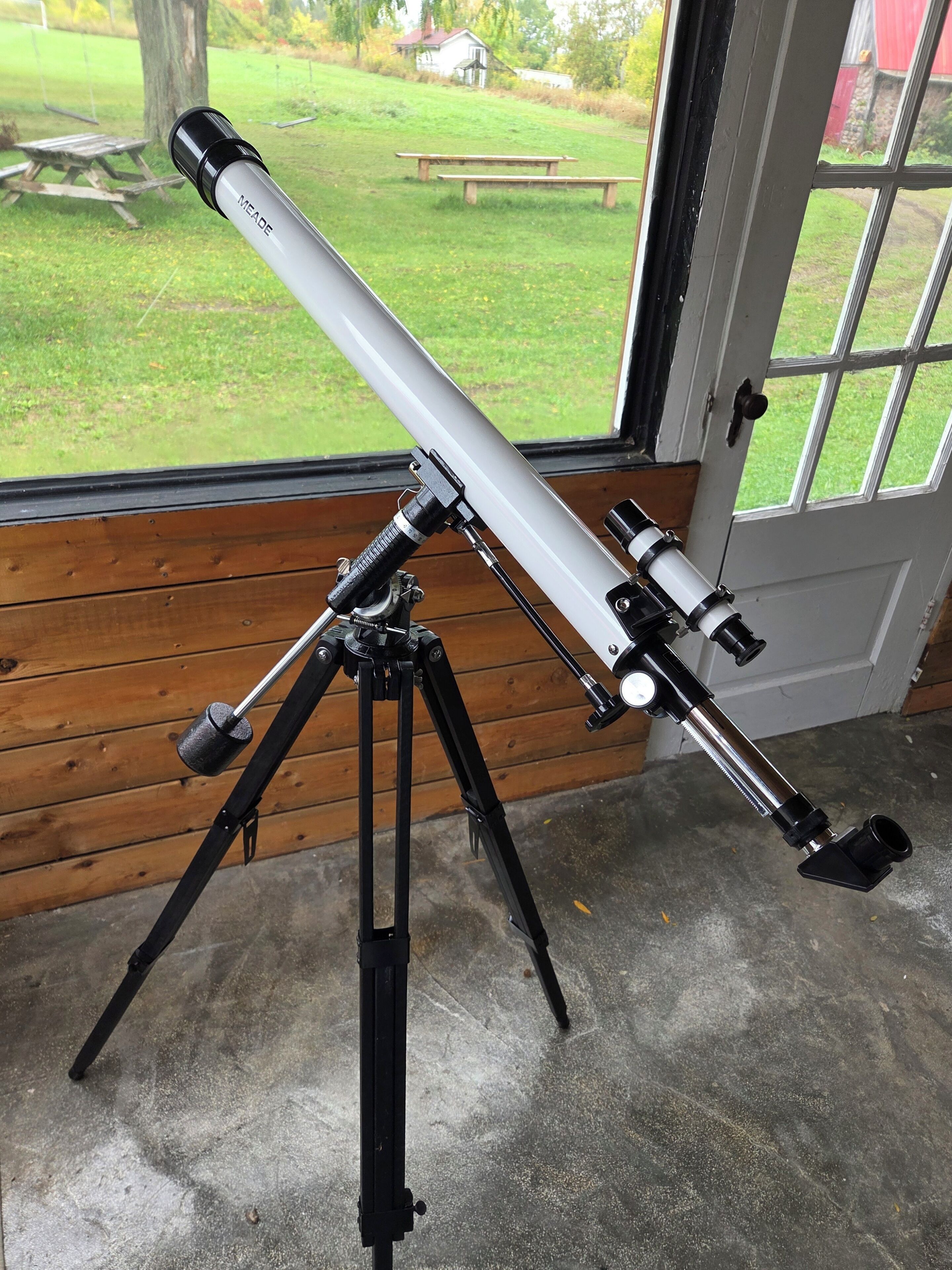 Un télescope monté sur un trépied, à l’intérieur d’une pièce dont un mur est en bois, et une porte donne sur un espace verdoyant équipé de tables de pique-nique.