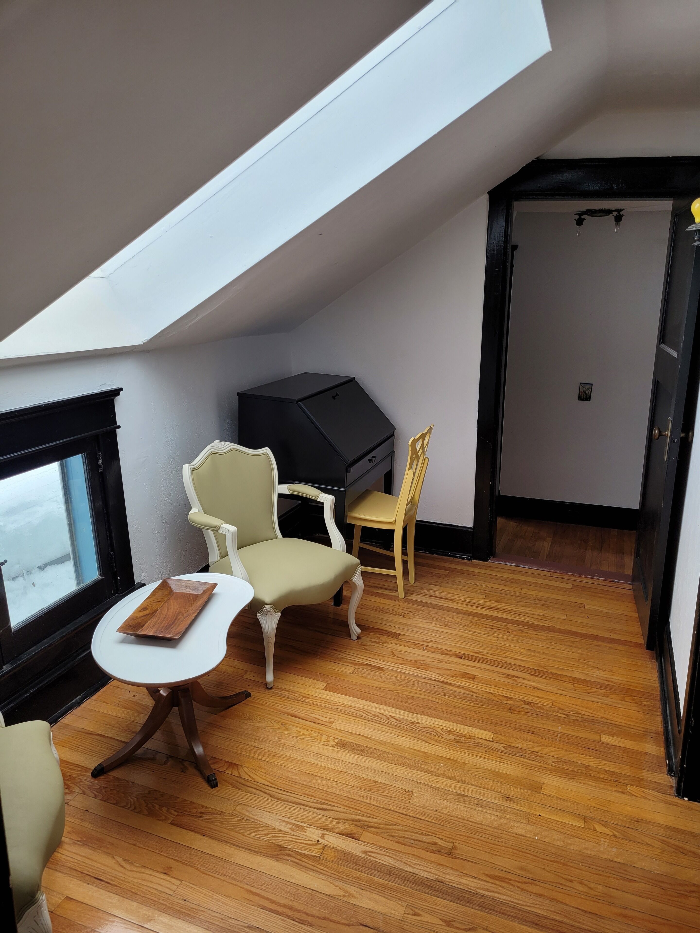 Une pièce avec un plafond en pente, un meuble noir, une table blanche, un fauteuil beige, une chaise jaune et un plancher en bois.