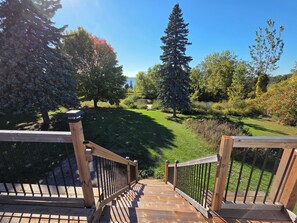 Une terrasse en bois avec une rambarde en métal, qui mène à une zone herbeuse agrémentée d’arbres et offrant une vue sur un ciel dégagé.