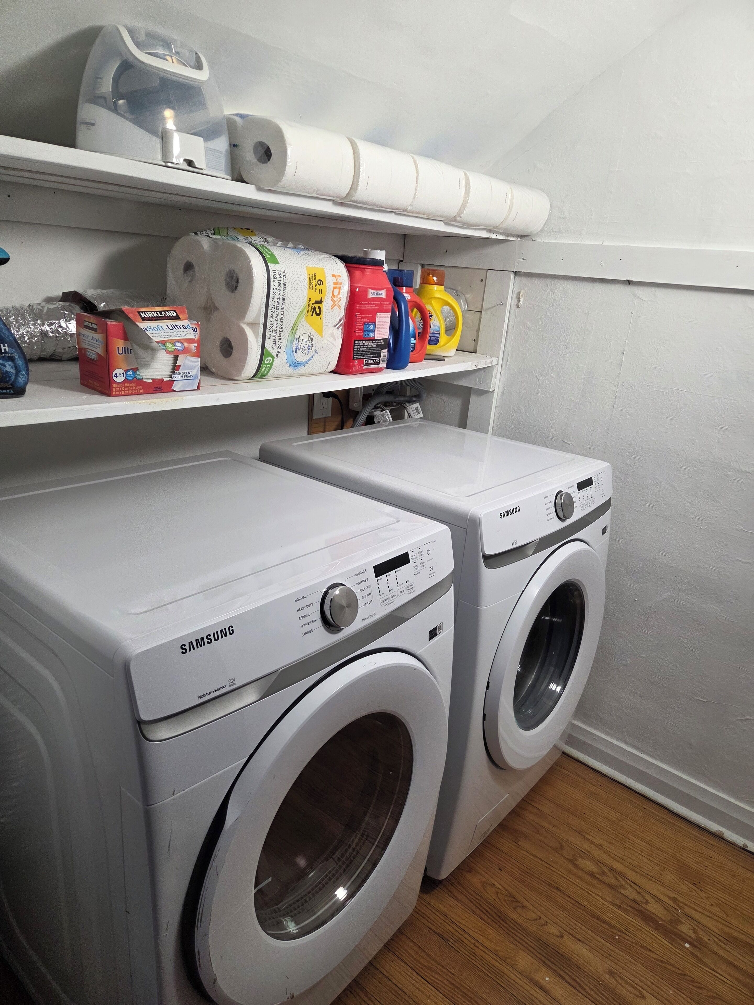 A laundry area with two Samsung washing machines, a shelf with various laundry supplies, and a white wall.