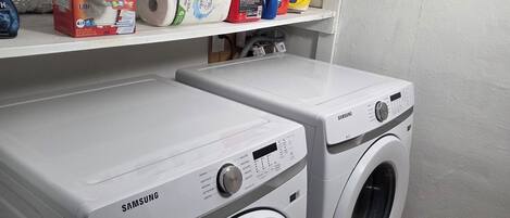 A laundry area with two Samsung washing machines, a shelf with various laundry supplies, and a white wall.