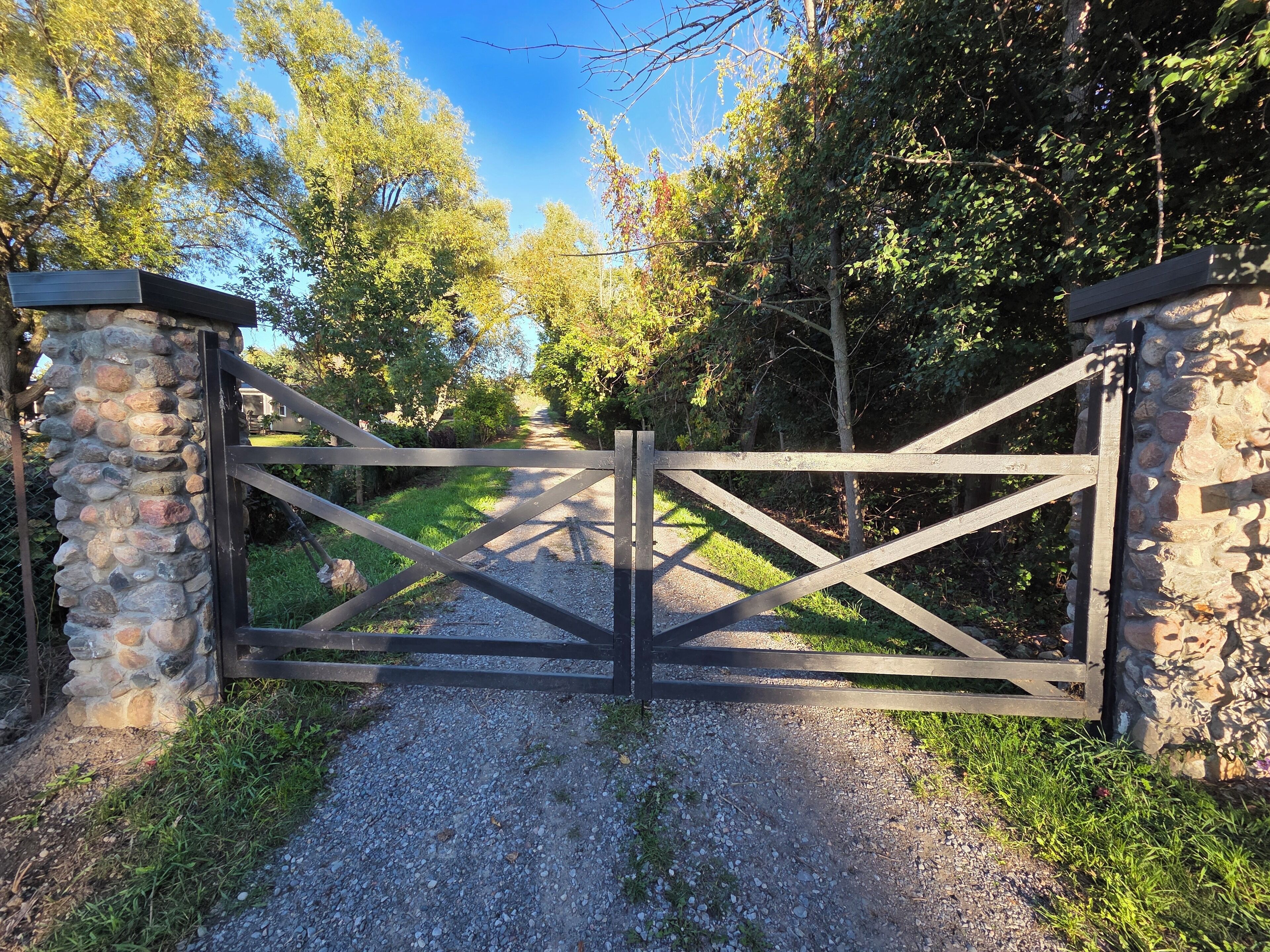 A rustic stone gate with a wooden crossbar, leading to a gravel path surrounded by greenery.