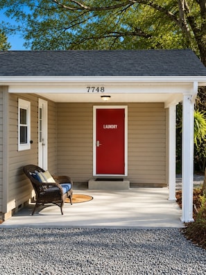 A house with a red door labeled 'Laundry,' a porch with a wicker chair, and a gravel driveway.