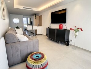 A modern living room with a grey sofa, a colorful ottoman, a black entertainment unit, and a flat-screen TV mounted on the wall.