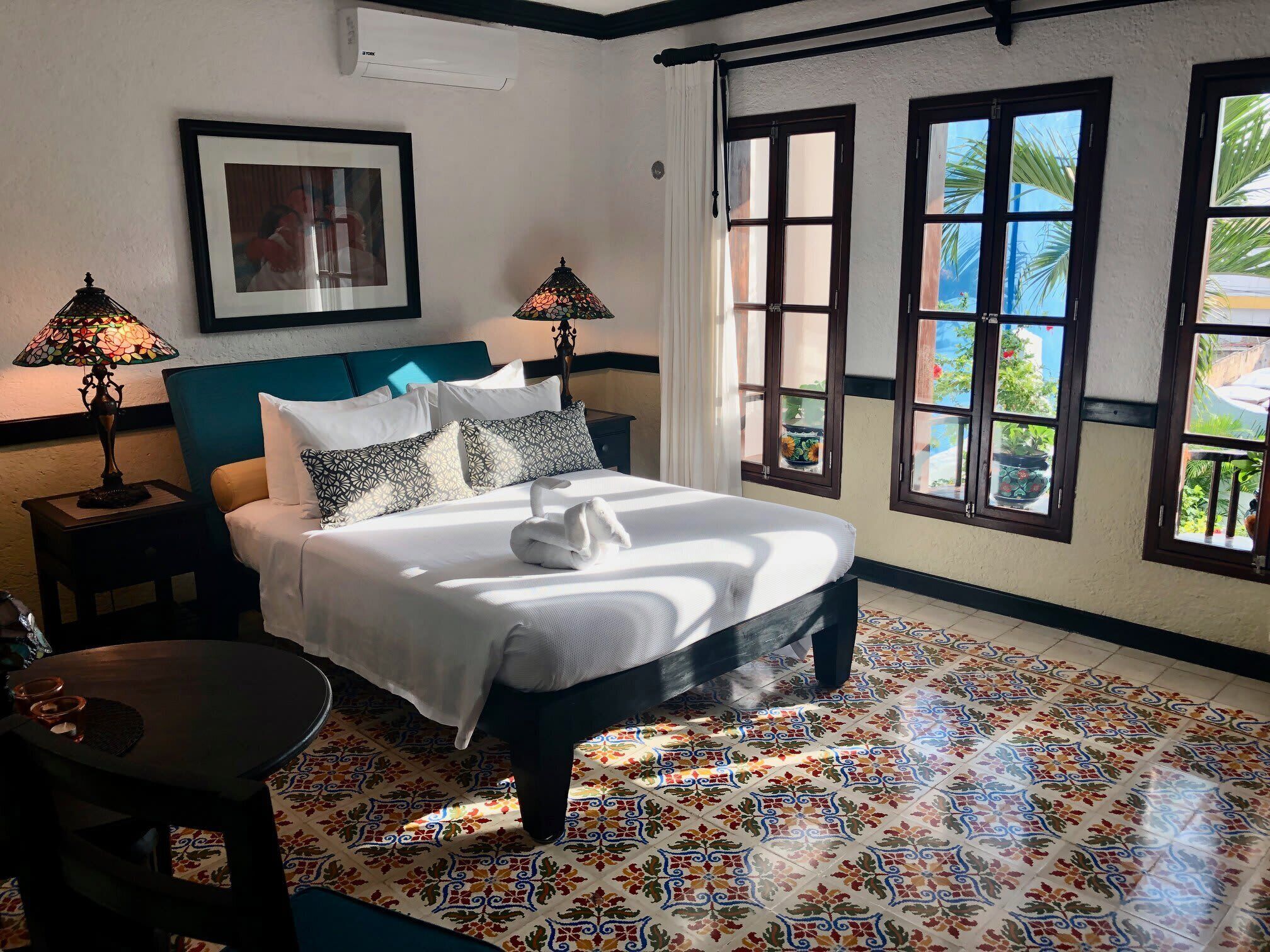 A neatly made bed with a white comforter and two pillows, a bedside table with a lamp, and a view of the outdoors through large windows.