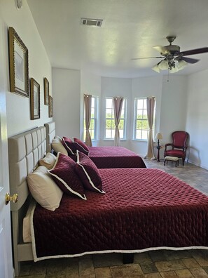 Ein Schlafzimmer mit zwei Betten, einem Deckenventilator und großen Fenstern mit Vorhängen.