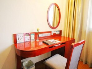 Une chambre d’hôtel avec un bureau en bois, un miroir, un petit vase avec une fleur, une poubelle et une chaise.