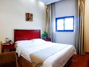 Une chambre d’hôtel avec une tête de lit rouge, un lit avec du linge de lit blanc, une table de chevet avec un téléphone et une plante, et une fenêtre avec des rideaux.