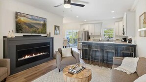 Una sala de estar moderna con chimenea, ventilador de techo y una zona de cocina con taburetes de bar.