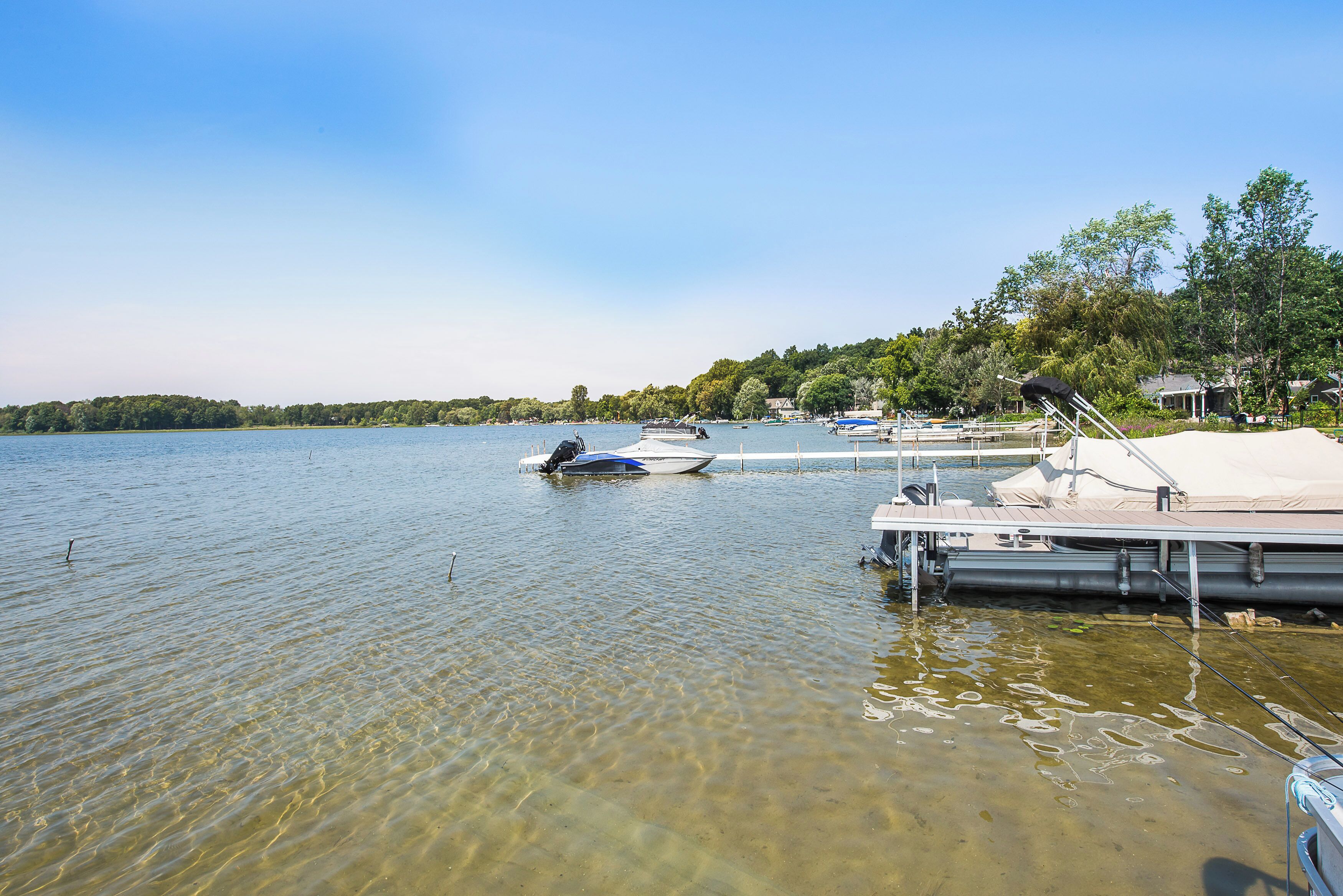 A calm lake with boats docked at a pier, surrounded by trees and a clear sky.