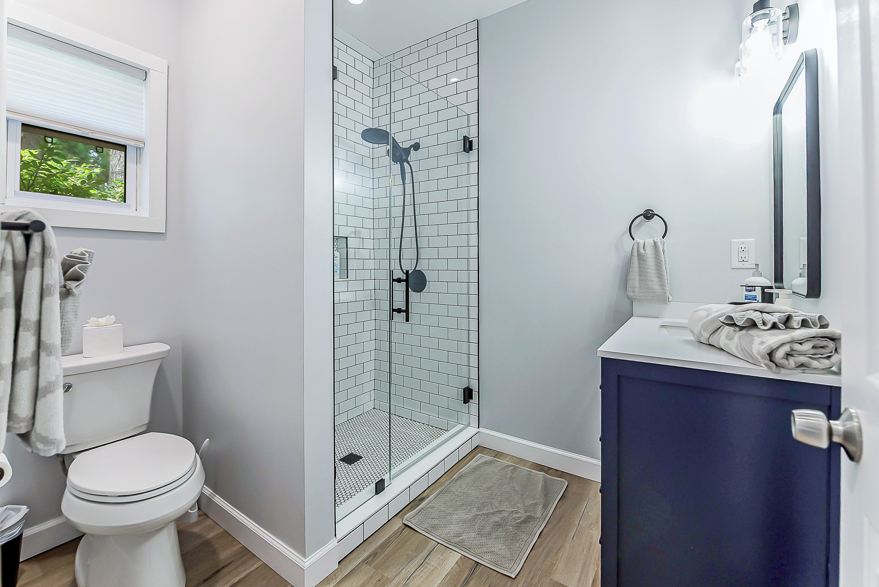 A bathroom with a glass shower enclosure, a toilet, a sink with a towel, and a window with a view of greenery.