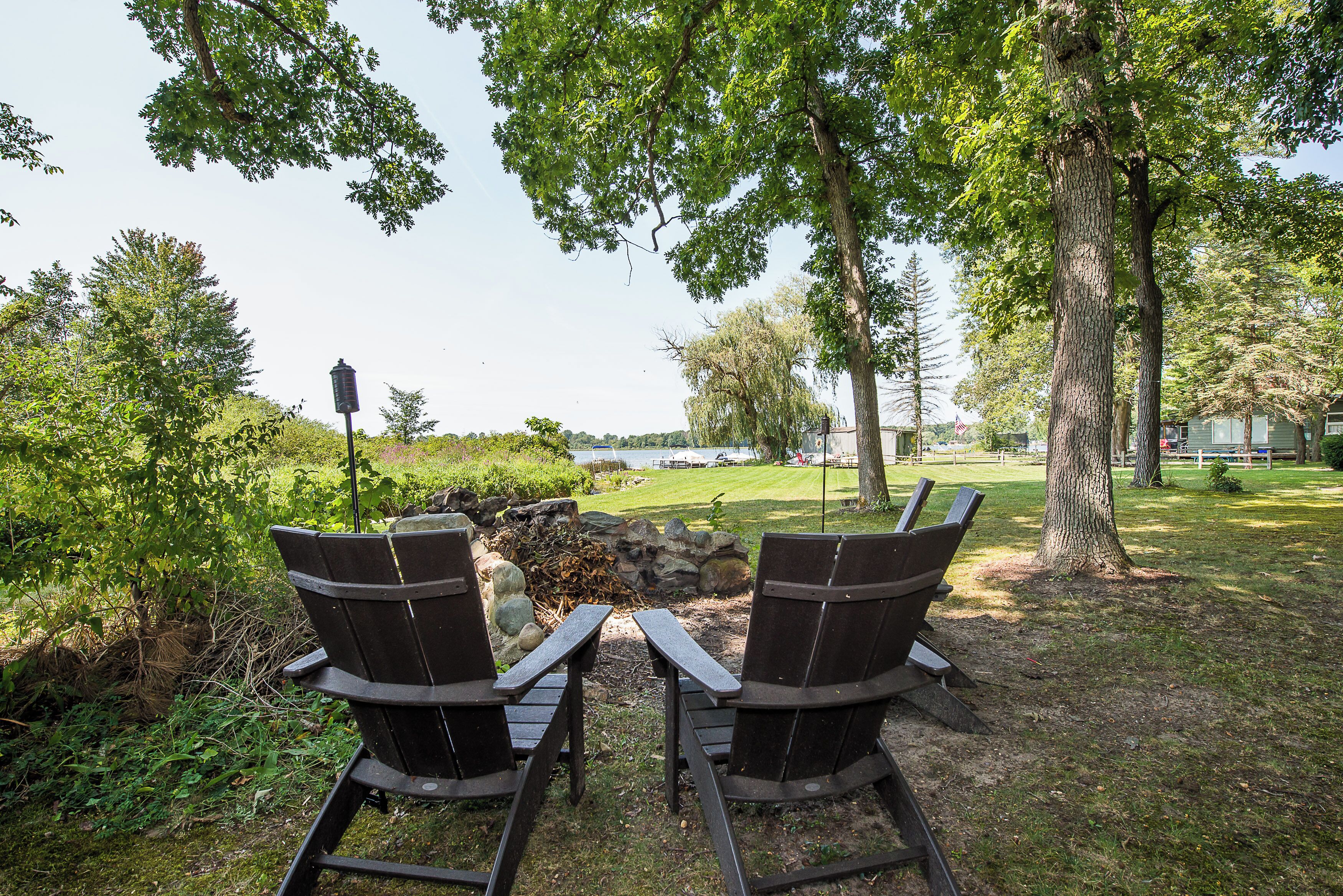 Two Adirondack chairs facing a fire pit with a view of a lake and a grassy area with trees.