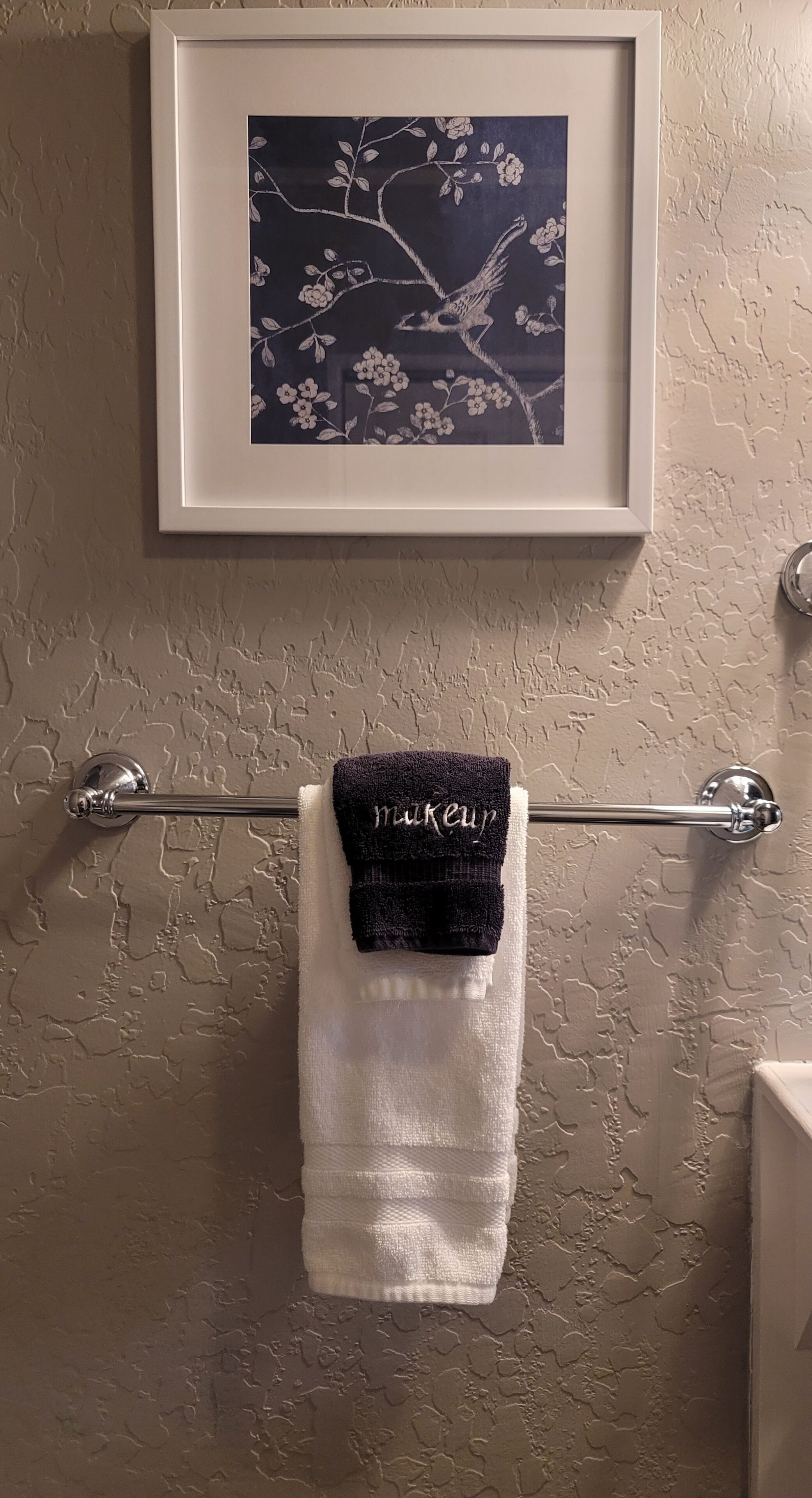 Un baño con una pared texturizada, una obra de arte enmarcada con pájaros y flores, y un perchero con una toalla blanca y una toalla negra.