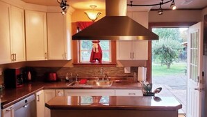 A modern kitchen with white cabinets, a stainless steel range hood, and a large window overlooking a garden. - Idyllic Cottage Memory Making with Pool & Outdoor Fireplace under the Gazebo, (Spokane Valley)