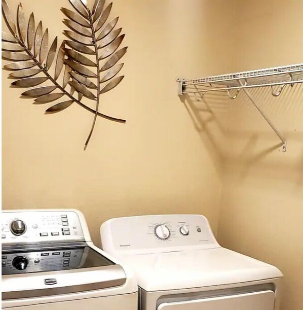 A laundry area with a white washer and dryer, a metal drying rack, and a decorative leaf wall art piece. - Eclectic Ranch Style Home - Sleeps 12 (Ocala)