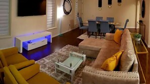A living room with a sofa, armchair, coffee table, and TV. There is a dining area with a table and chairs.