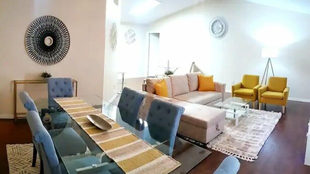 A modern living room with a glass dining table, blue chairs, a beige sofa with orange cushions, and yellow armchairs.