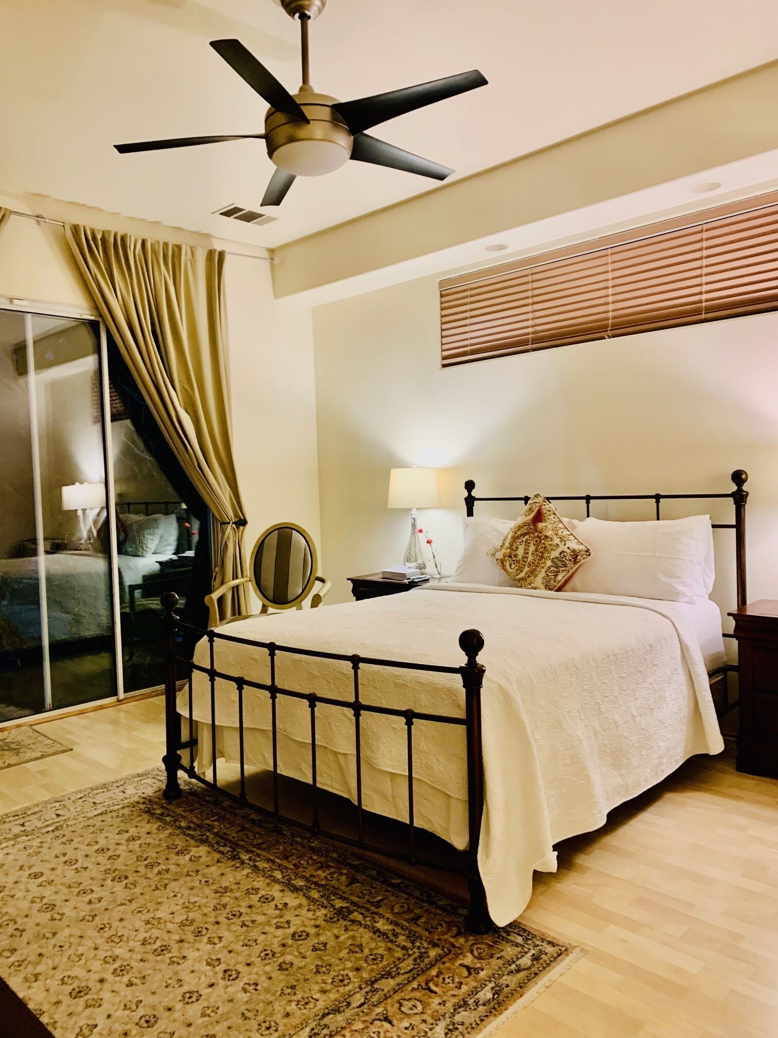 Un dormitorio con cama, ventilador de techo, ventana con persianas, mesita de noche con lámpara y una silla.