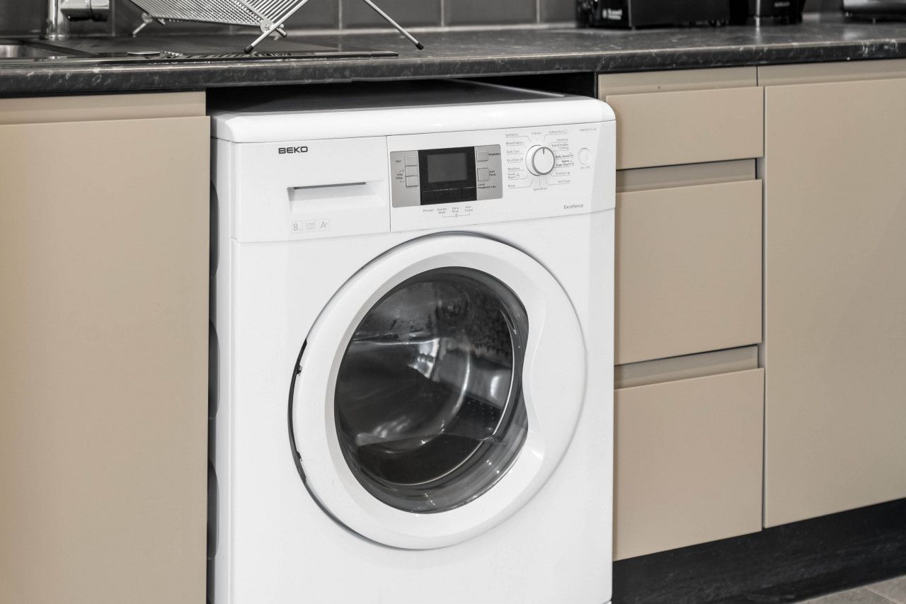 Un lave-linge BEKO à chargement frontal, de couleur blanche, se trouve dans une cuisine avec des armoires de couleur beige et un comptoir noir.
